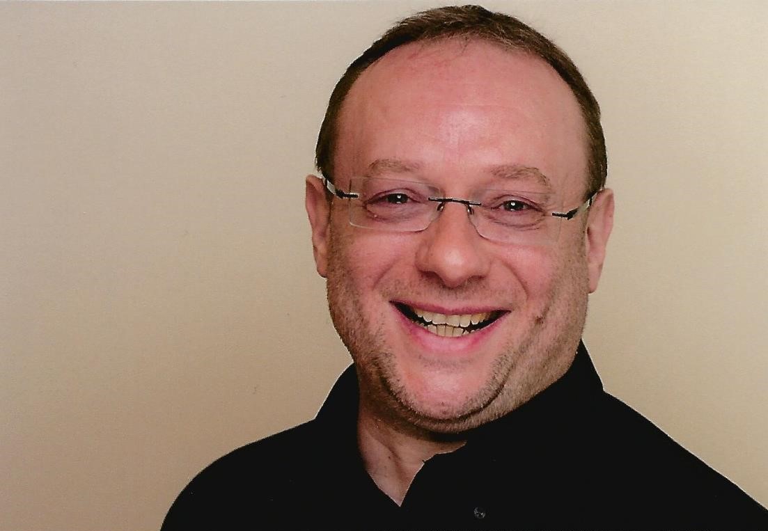 Ein Mann mit kurzen Haaren, 3-Tage-Bart und Brille lacht in die Kamera.